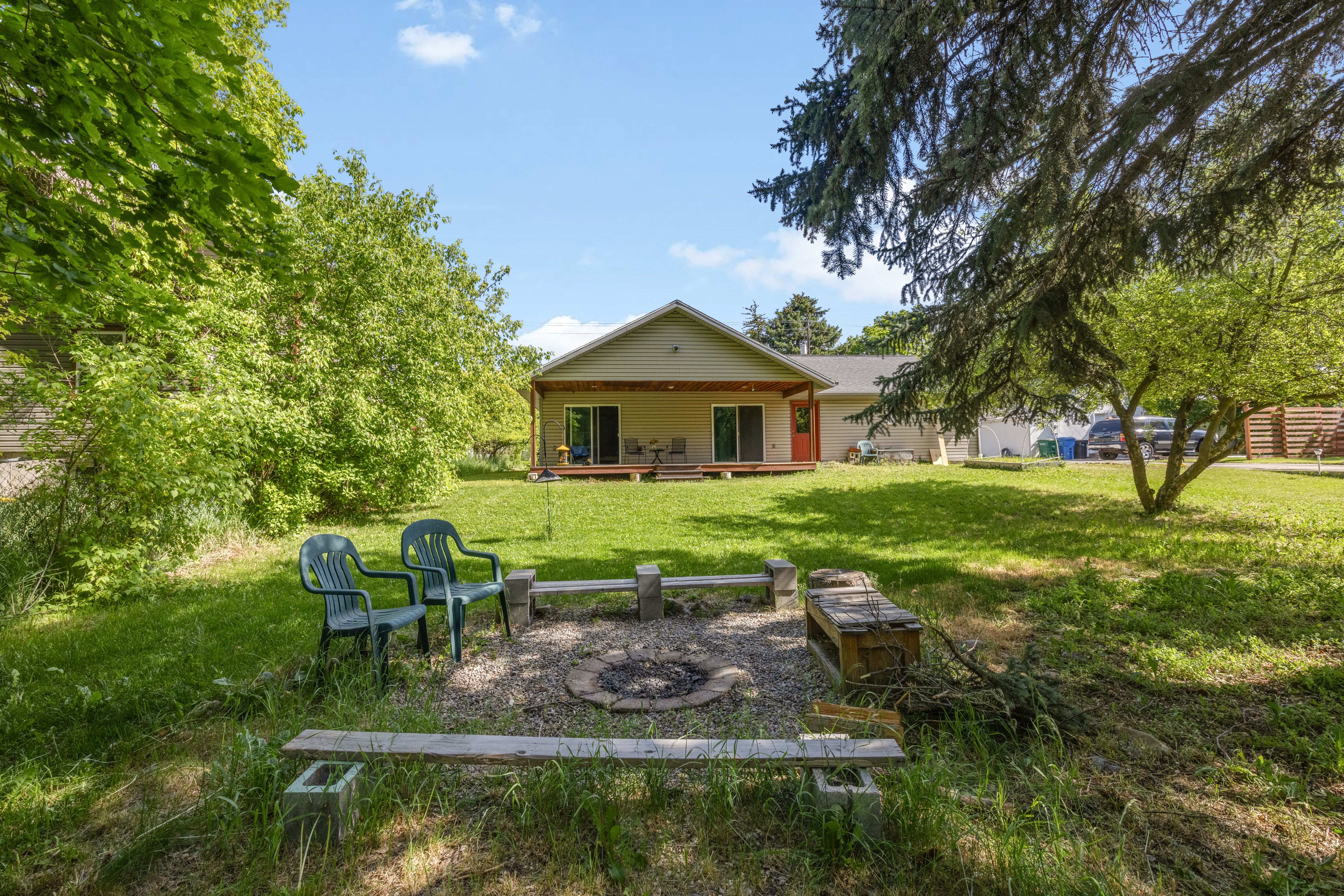 Spacious yard featuring a built-in fire pit with stone-block seating, expansive lawn, mature trees, and a back deck with sliding glass doors