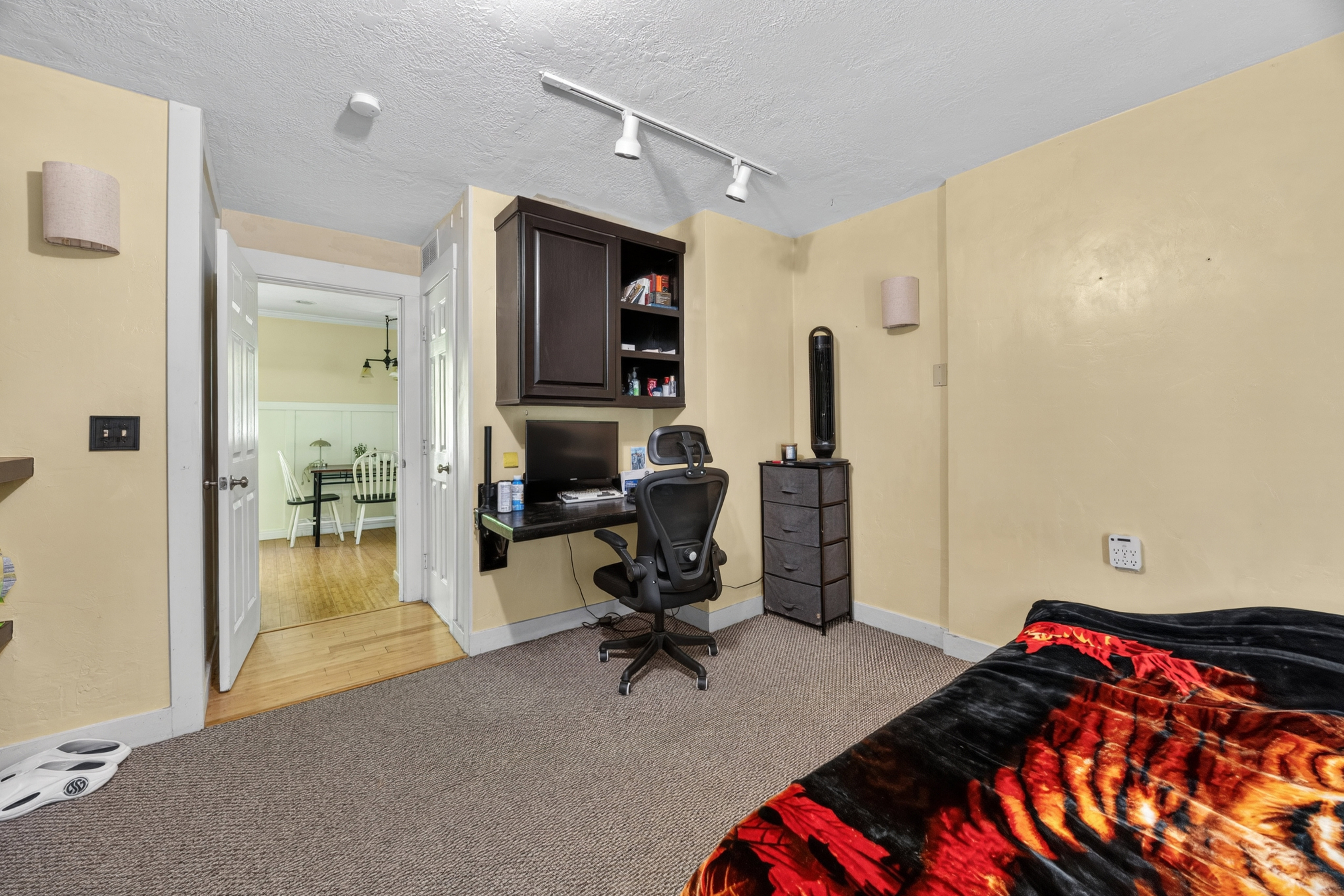 Carpeted room featuring a built-in dark wood desk with shelving, track lighting, and a doorway opening to a room with wood flooring and wainscoting