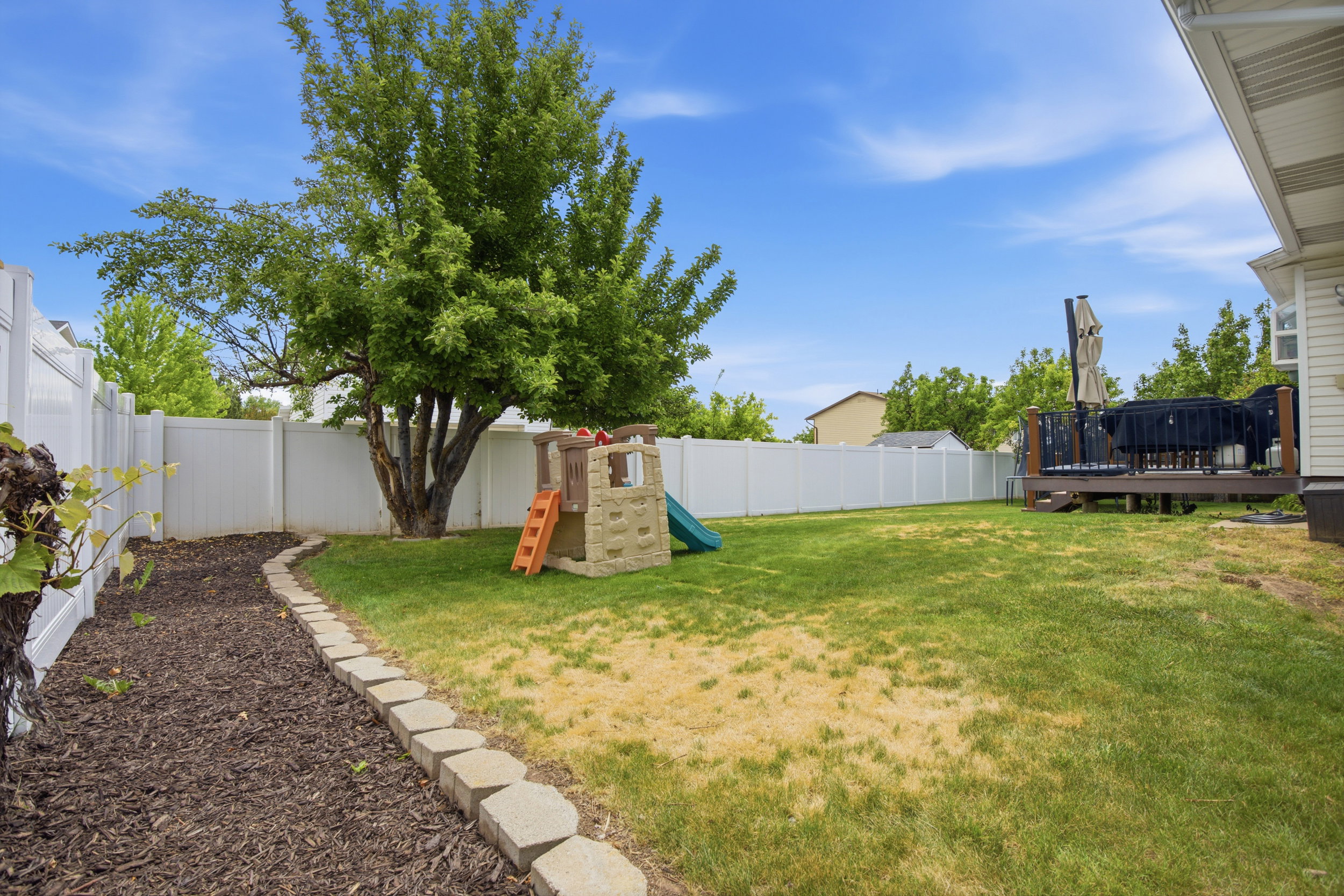 Expansive backyard featuring a full vinyl privacy fence, mature tree, multi-level wood deck, and a defined planting bed with stone edging