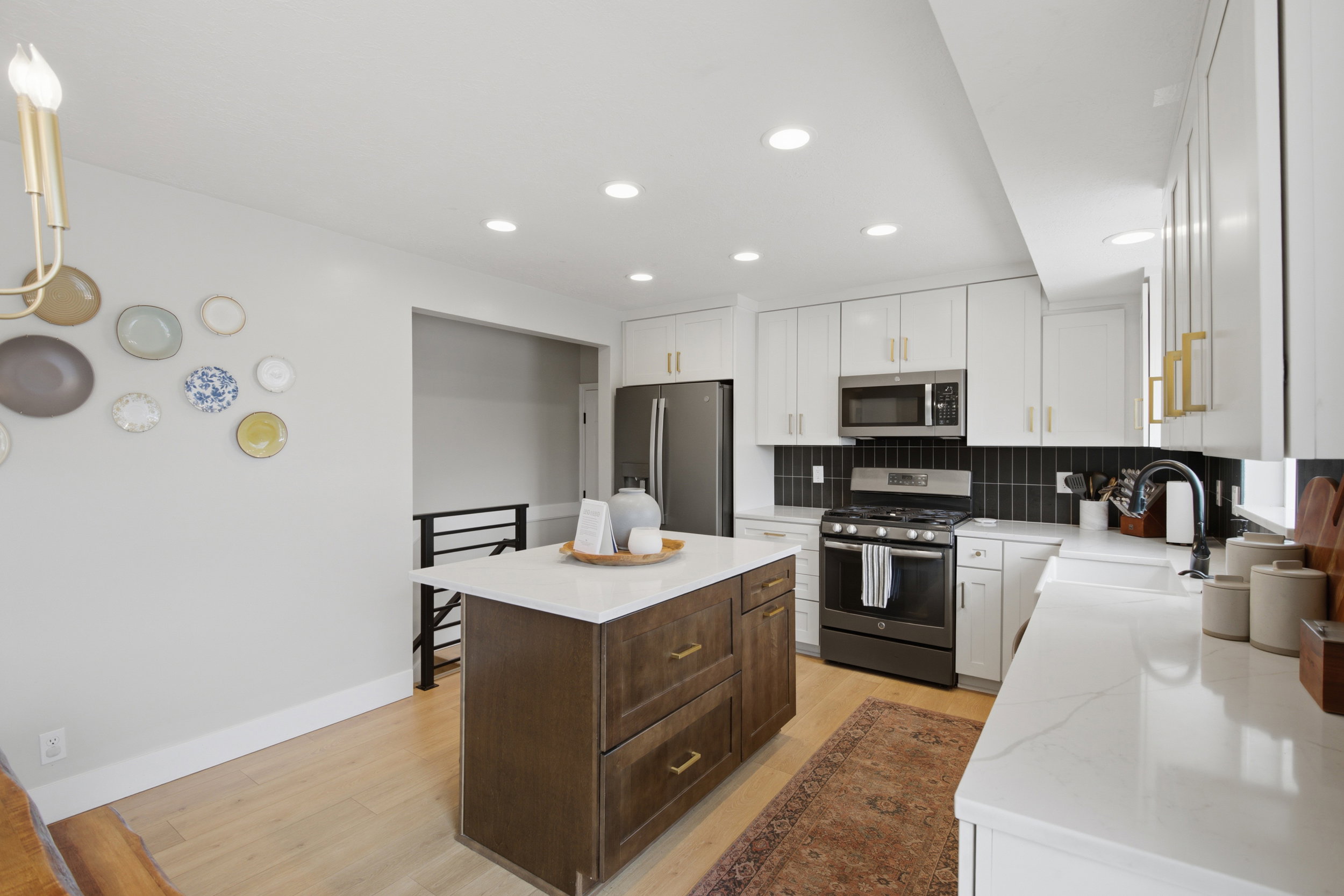 Modern kitchen featuring a central island with a light-toned countertop, white cabinetry with gold-toned hardware, a dark subway tile backsplash, stainless steel appliances, and light wood-finish flooring