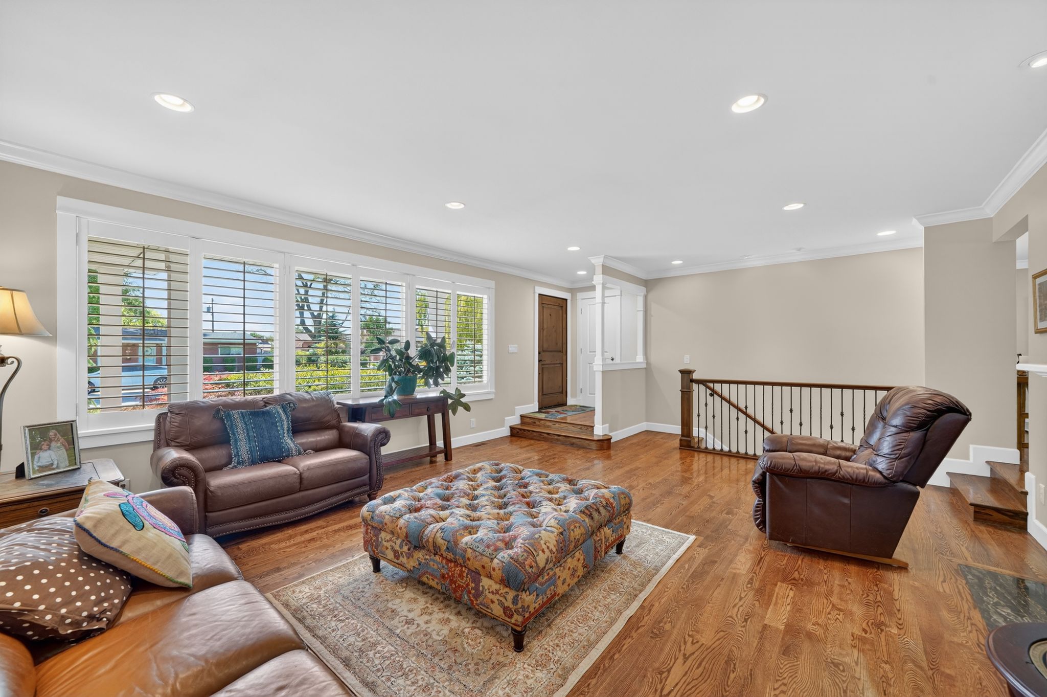 Spacious living area featuring polished wood flooring, recessed lighting, white crown molding, large windows with white louvered shutters, and a wooden handrail with black metal balusters