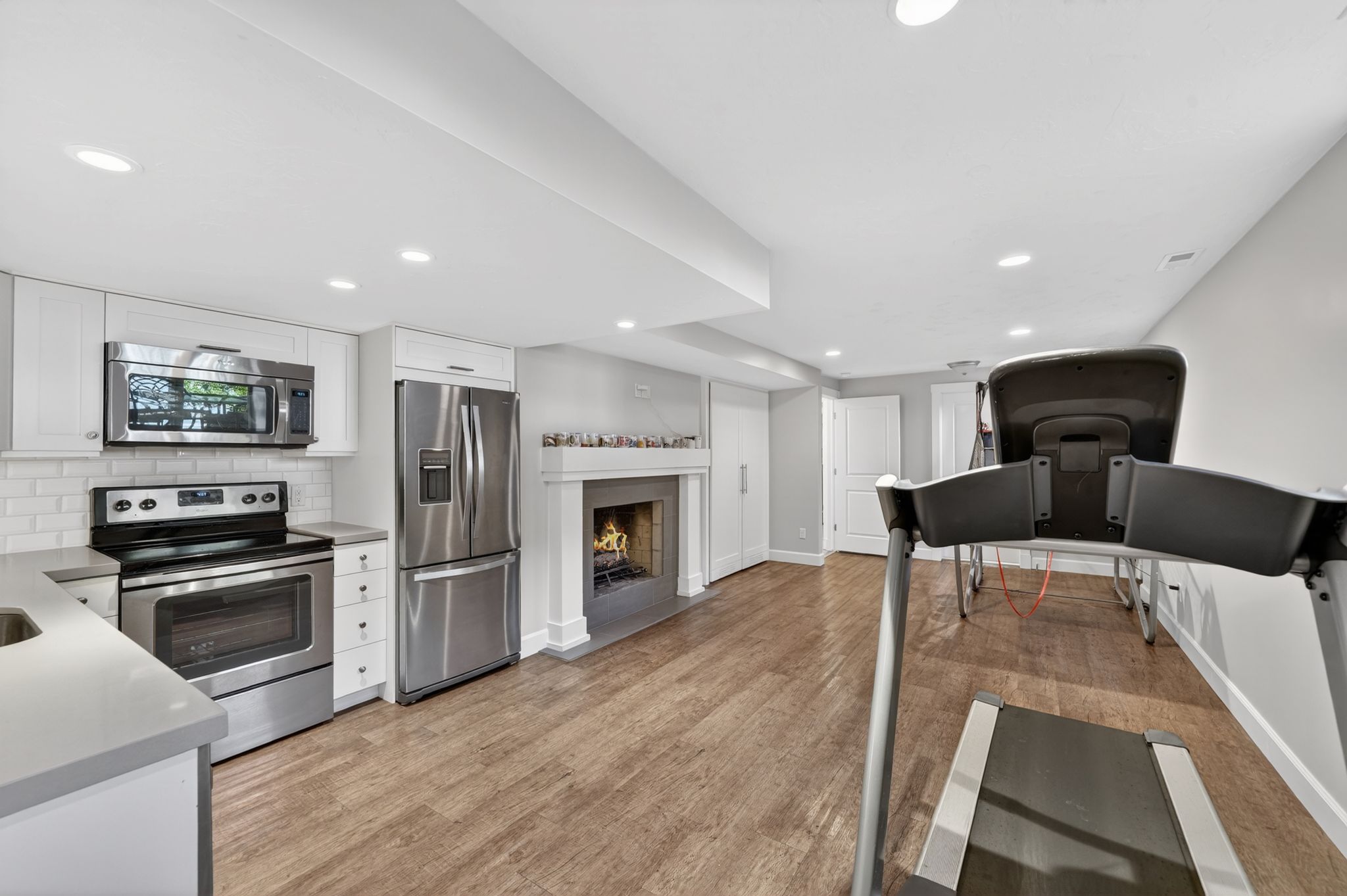 Wood-finish flooring throughout a living area featuring a built-in fireplace with a stone hearth, white mantel, and integrated kitchen cabinetry with stainless steel appliances