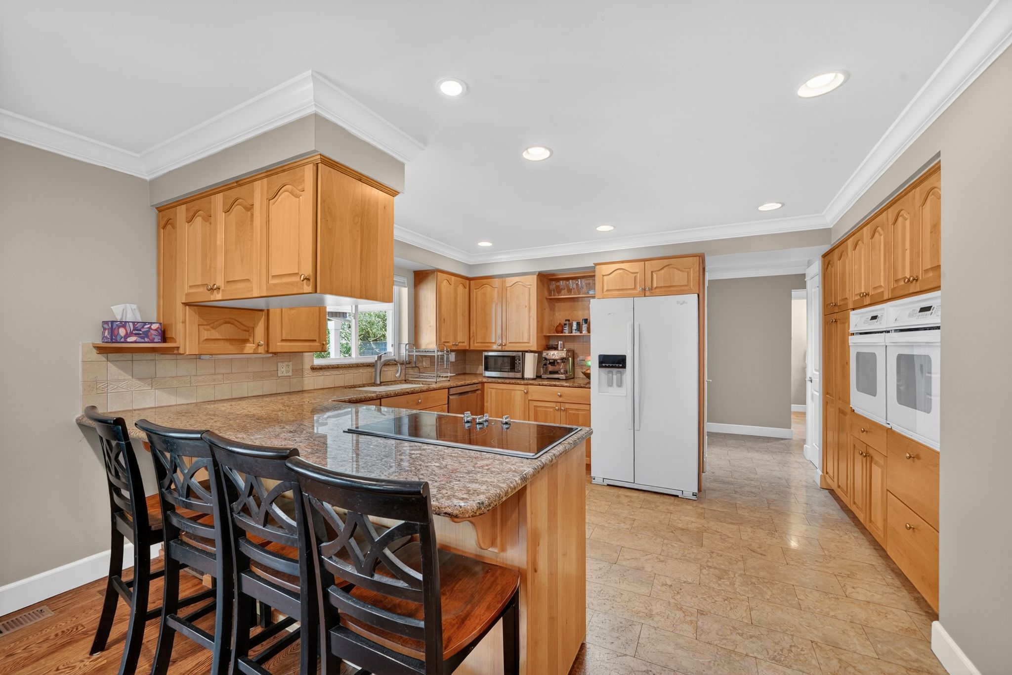 Spacious kitchen featuring light wood cabinetry, stone countertops, a breakfast bar with built-in sink, recessed lighting, and dual wall ovens