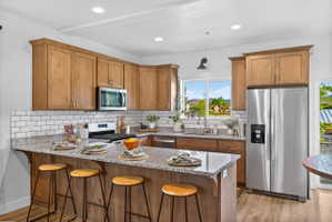 Kitchen featuring wood-finish cabinetry, stainless steel appliances, speckled stone countertops, a subway tile backsplash, and an island with bar seating