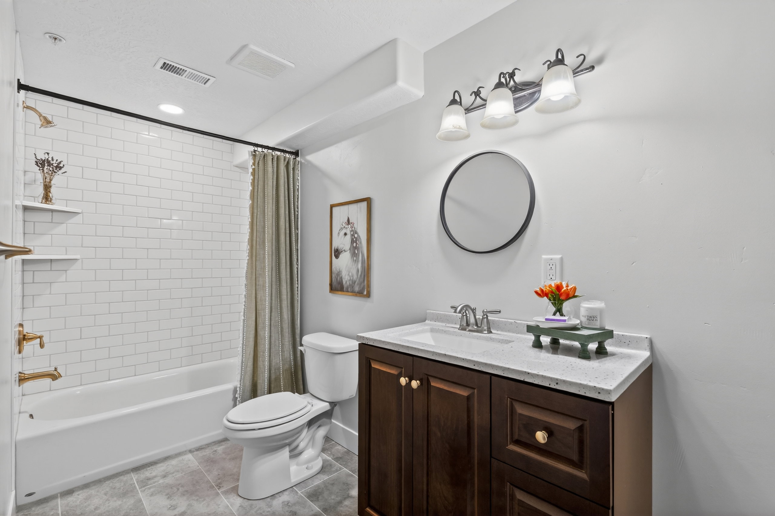 Bathroom featuring a white subway tile shower surround with gold-finish fixtures, a dark wood-finish vanity with a light-colored countertop, and a round wall mirror