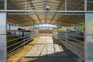 Covered equestrian facility featuring a metal roof structure, open-air stalls with metal bar fencing, a central concrete walkway, and overhead lighting fixtures