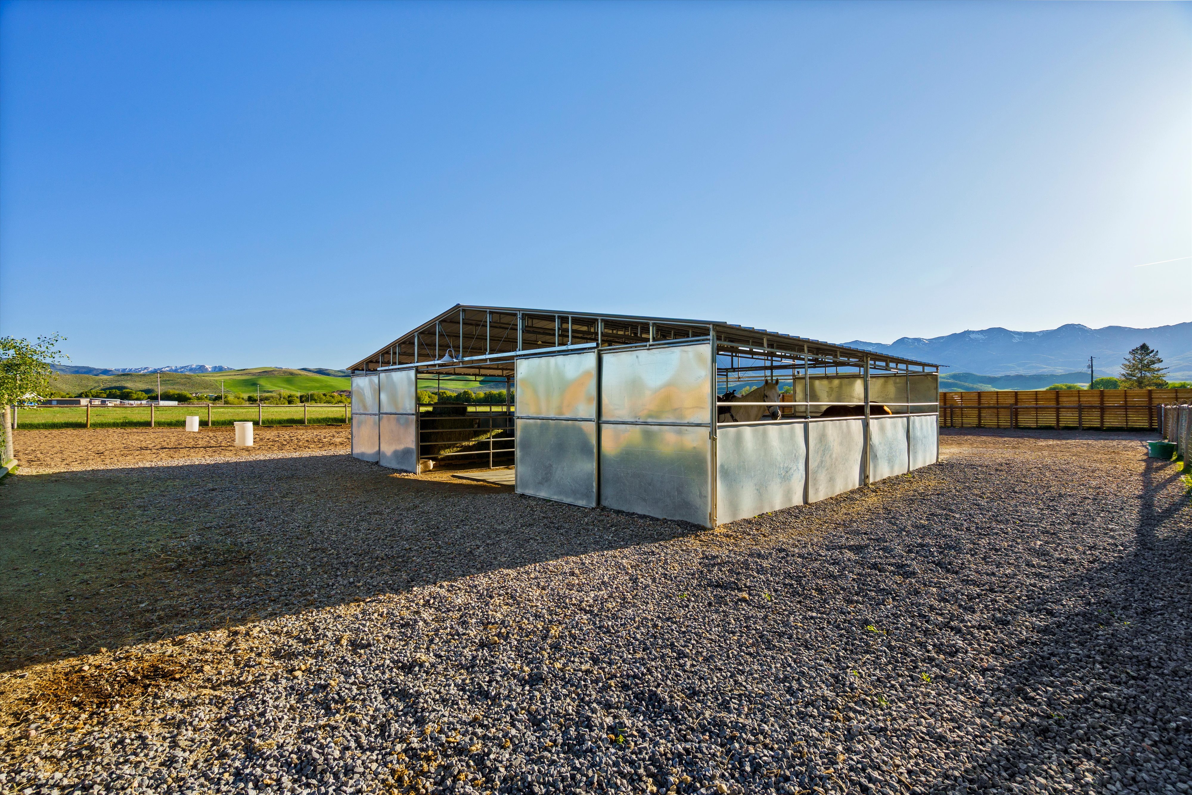 Open-air metal barn structure with multiple stalls, a gravel-surfaced paddock, and an adjacent dirt arena