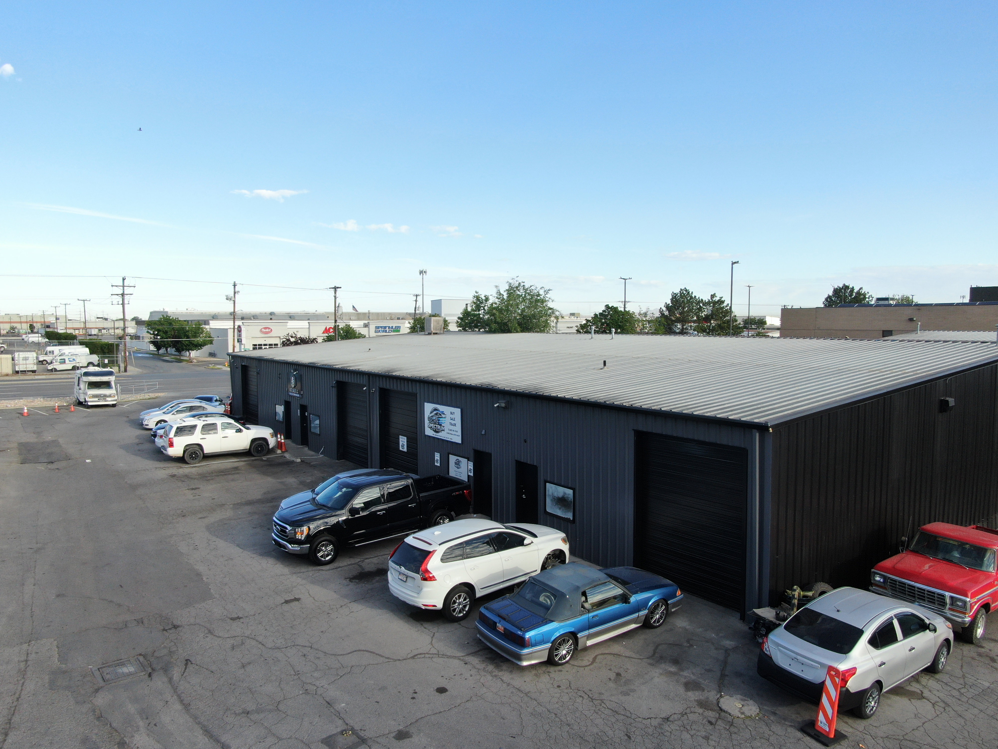 Industrial building featuring a dark corrugated metal exterior, multiple roll-up garage doors, a light-colored metal roof, and a paved lot