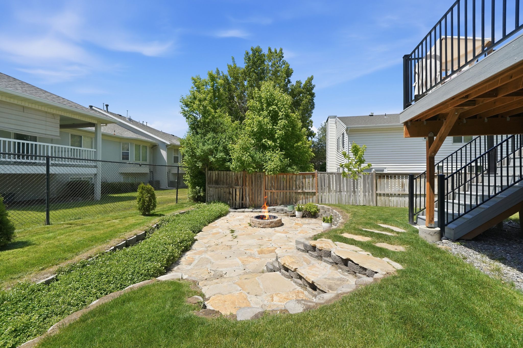 Expansive backyard featuring a natural stone patio with a built-in fire pit, stone-step pathways, a raised wood deck with black metal railings, and a fenced perimeter