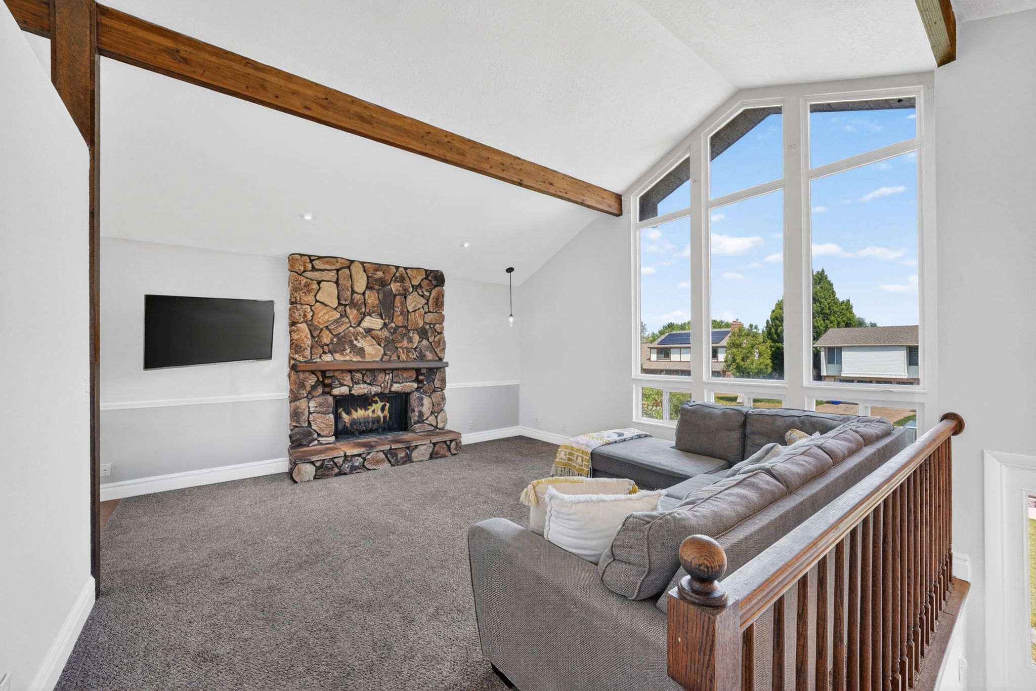 Vaulted ceiling living area featuring a stone fireplace, exposed wood beams, large picture windows, and carpet flooring
