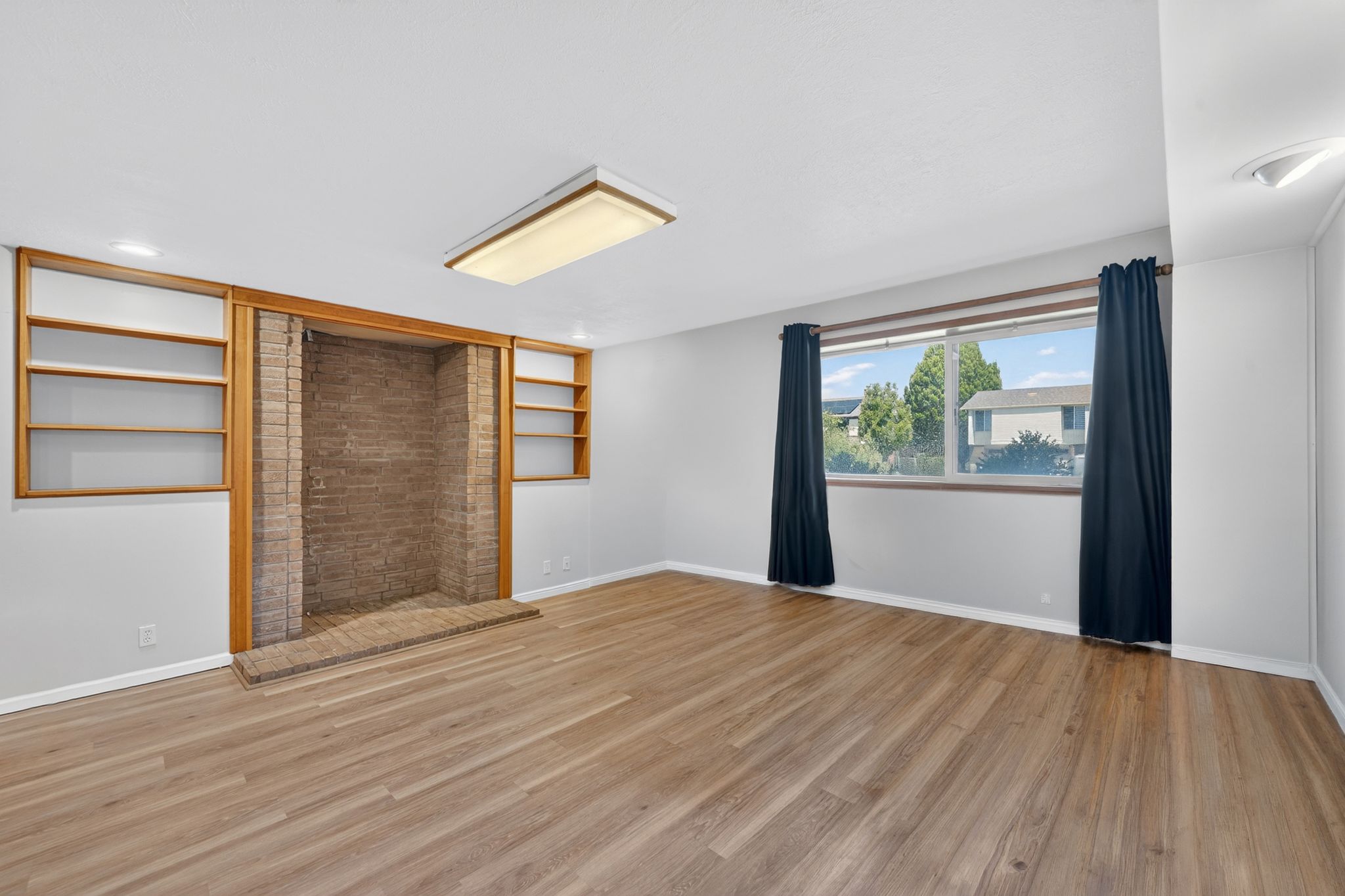 Spacious room featuring wood-finish flooring, a brick fireplace surround with hearth, built-in shelving, and a large window providing natural light