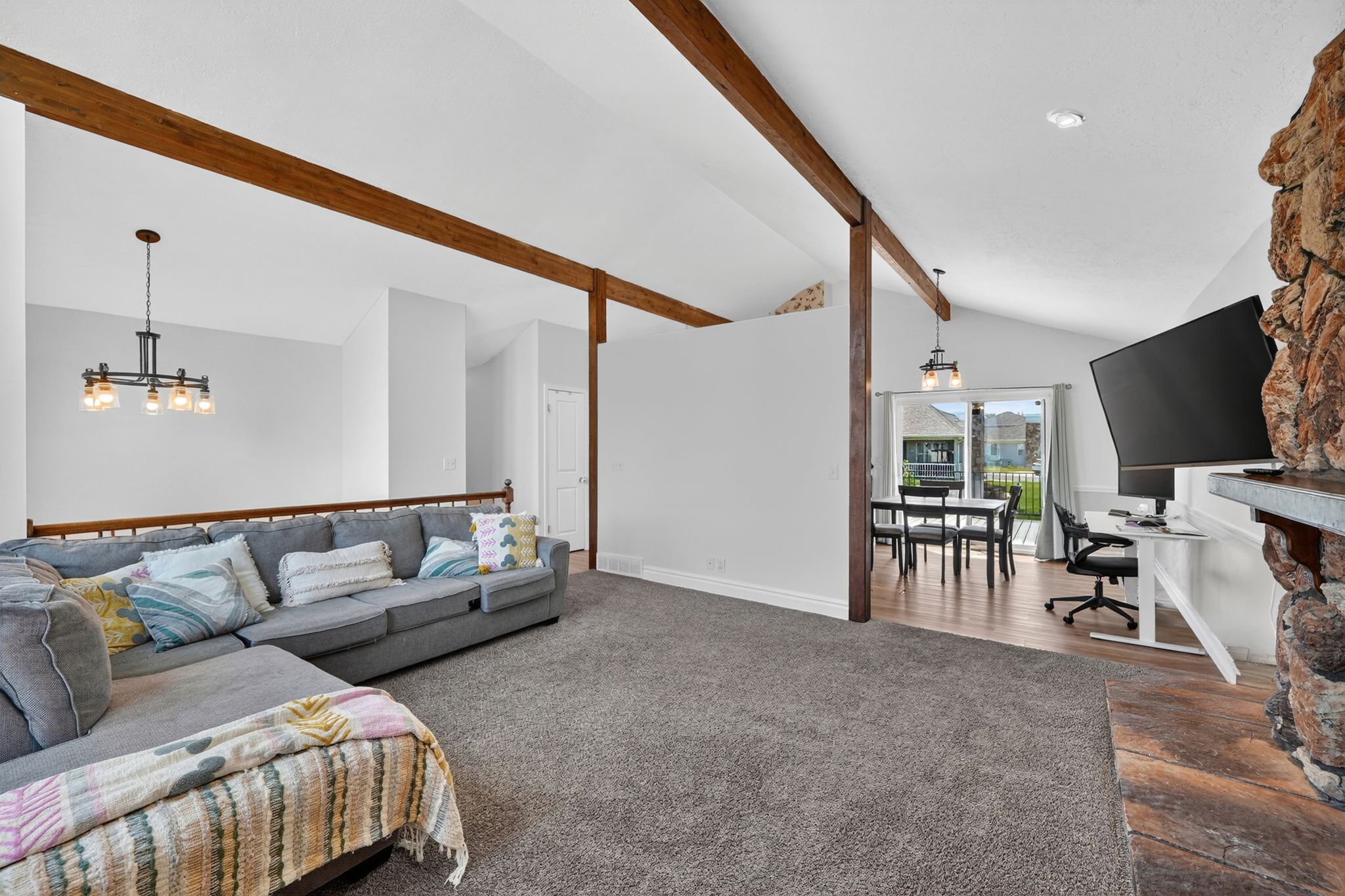 Open-concept living space featuring vaulted ceilings with exposed wood beams, a stone-clad fireplace, and gray carpeting