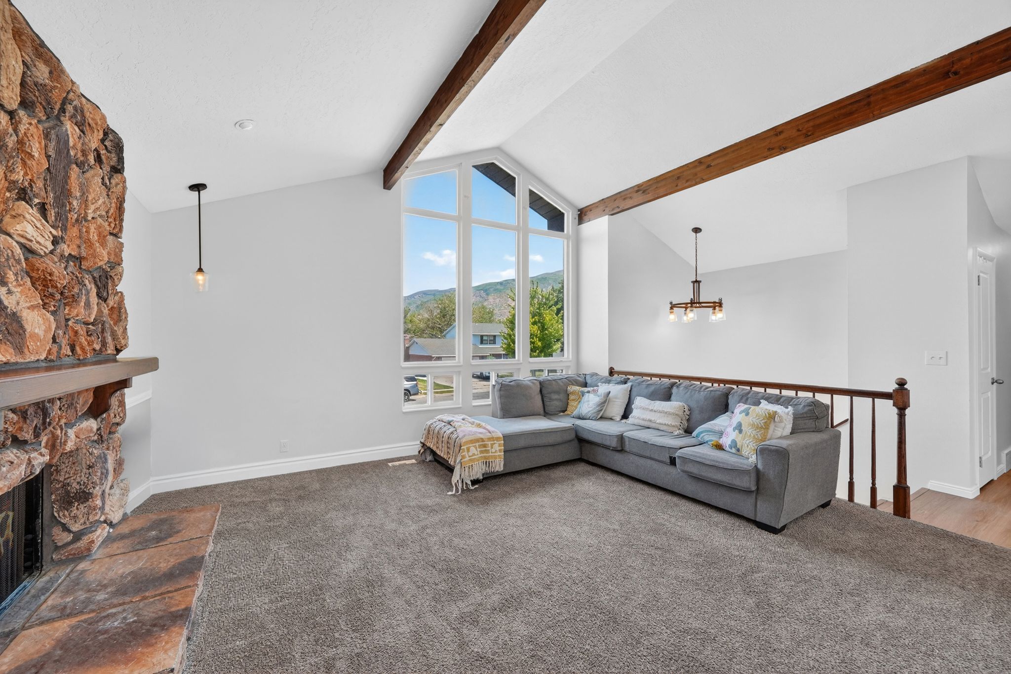 Vaulted ceiling with exposed wood beams, stone fireplace with mantel, oversized windows, and light gray wall finishes