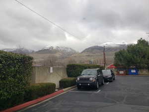 Paved parking area framed by manicured shrubbery and a solid wall, with distant snow-capped mountains under a cloudy sky