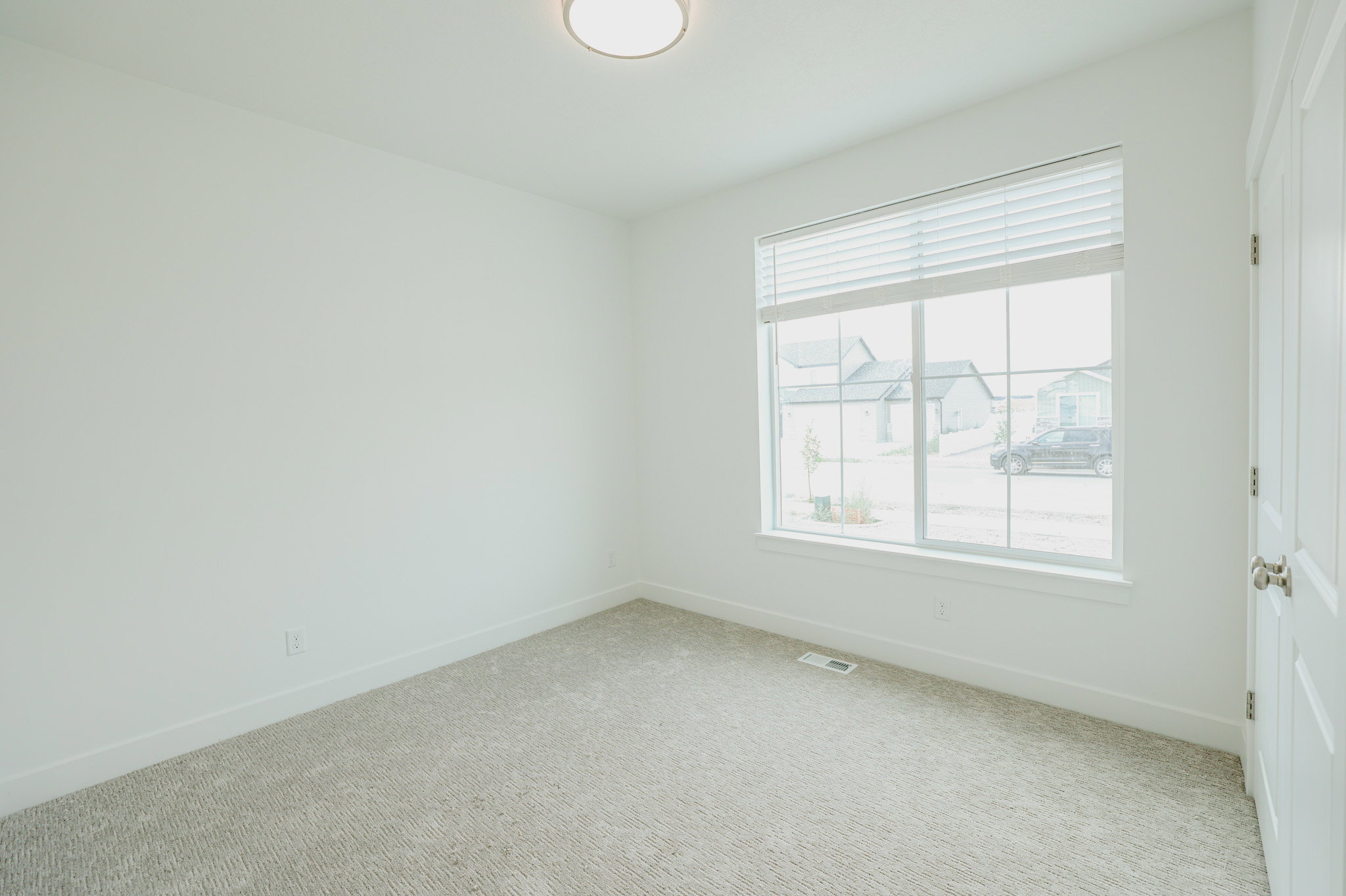 Bright room featuring a large window with blinds, neutral carpeting, a white paneled door, and a flush-mount ceiling light fixture