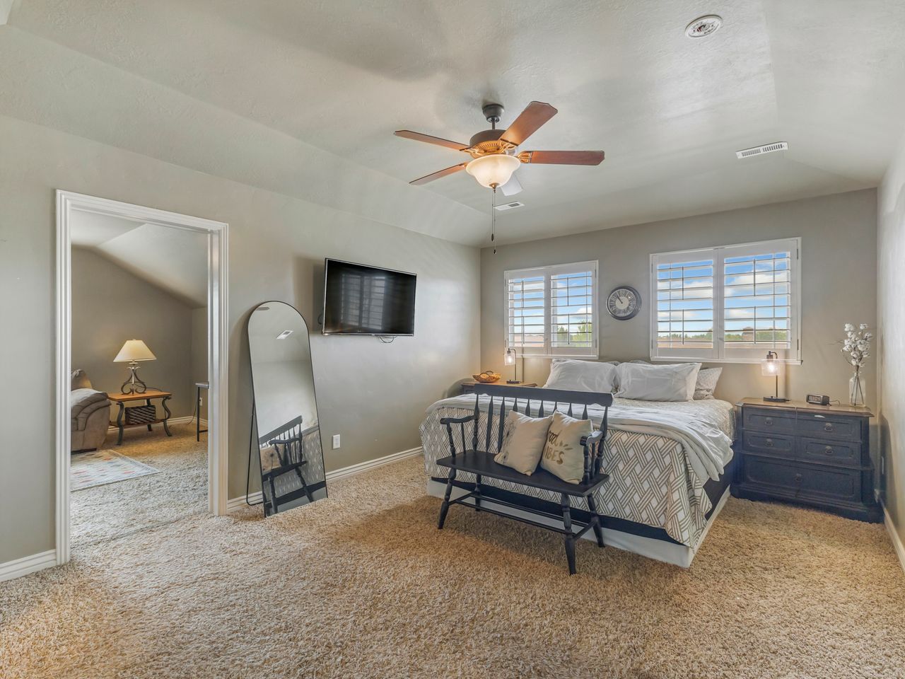 Carpeted room featuring a ceiling fan, two windows with white plantation shutters, and an open doorway revealing another carpeted room