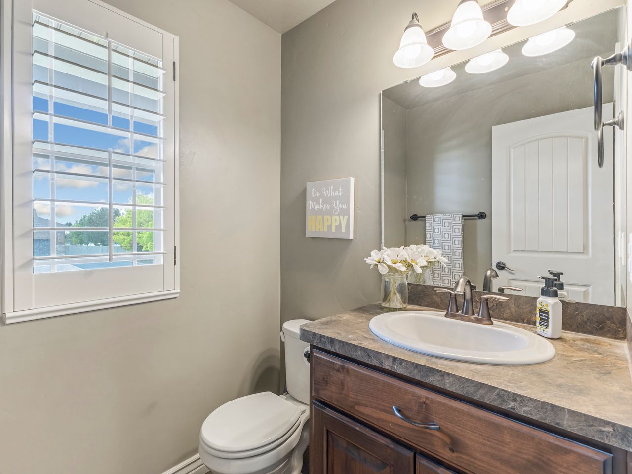Bathroom featuring a window with white louvered shutters, a dark wood-finish vanity with an integrated sink, a neutral-toned countertop, and a vanity mirror with an overhead light fixture