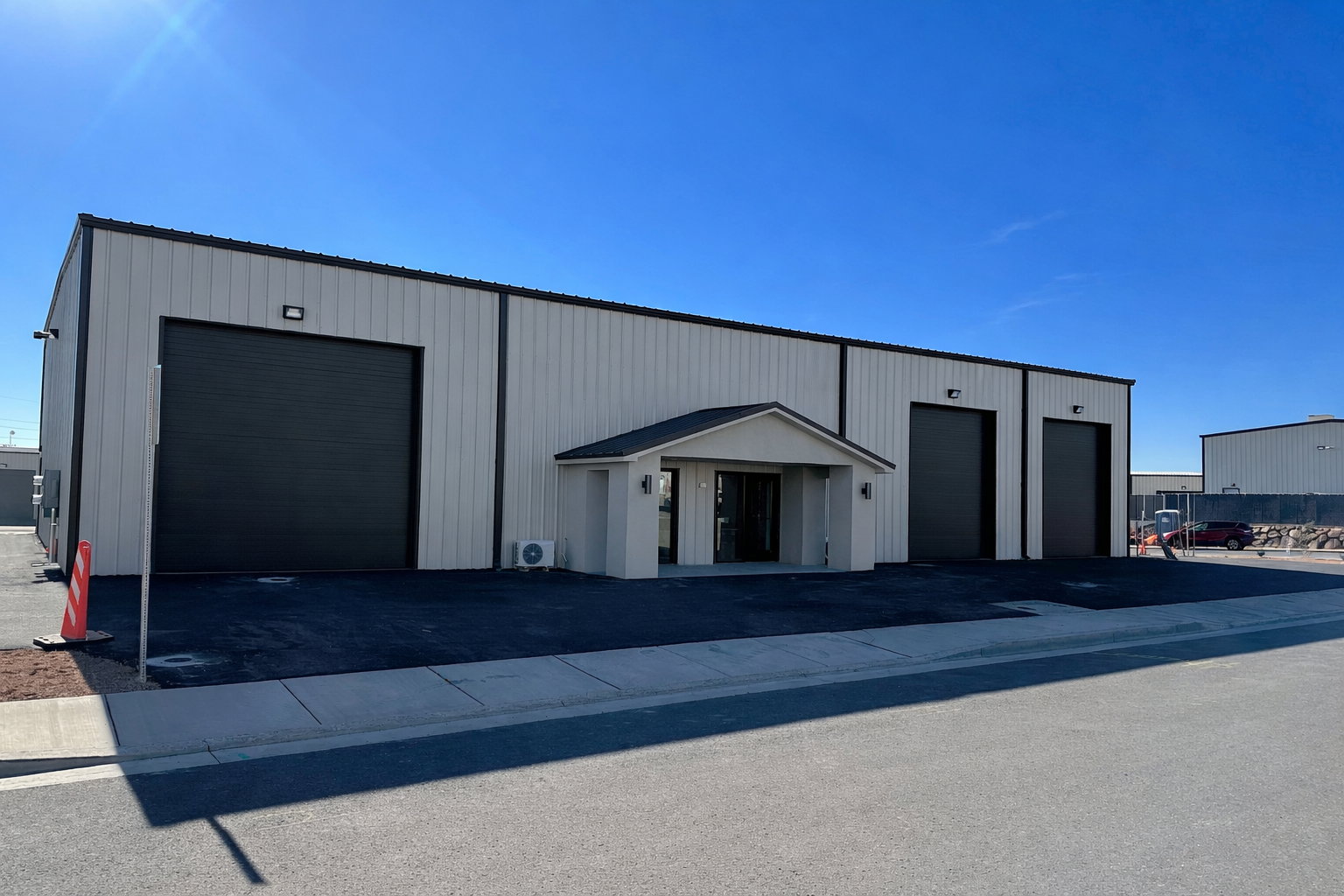 Commercial building featuring a light-colored metal exterior with black trim, three dark-colored roll-up doors, and an entry vestibule with an awning