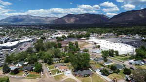 Expansive aerial perspective showcasing a residential area with varied architectural styles, mature trees, commercial buildings, and distant mountains under a blue sky with clouds
