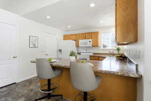 Kitchen featuring a breakfast bar with stone-finish countertops, wood cabinetry, white appliances, recessed lighting, and resilient flooring