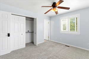 Bedroom featuring light blue walls, gray carpeting, a ceiling fan with light fixture, and a window with white plantation shutters
