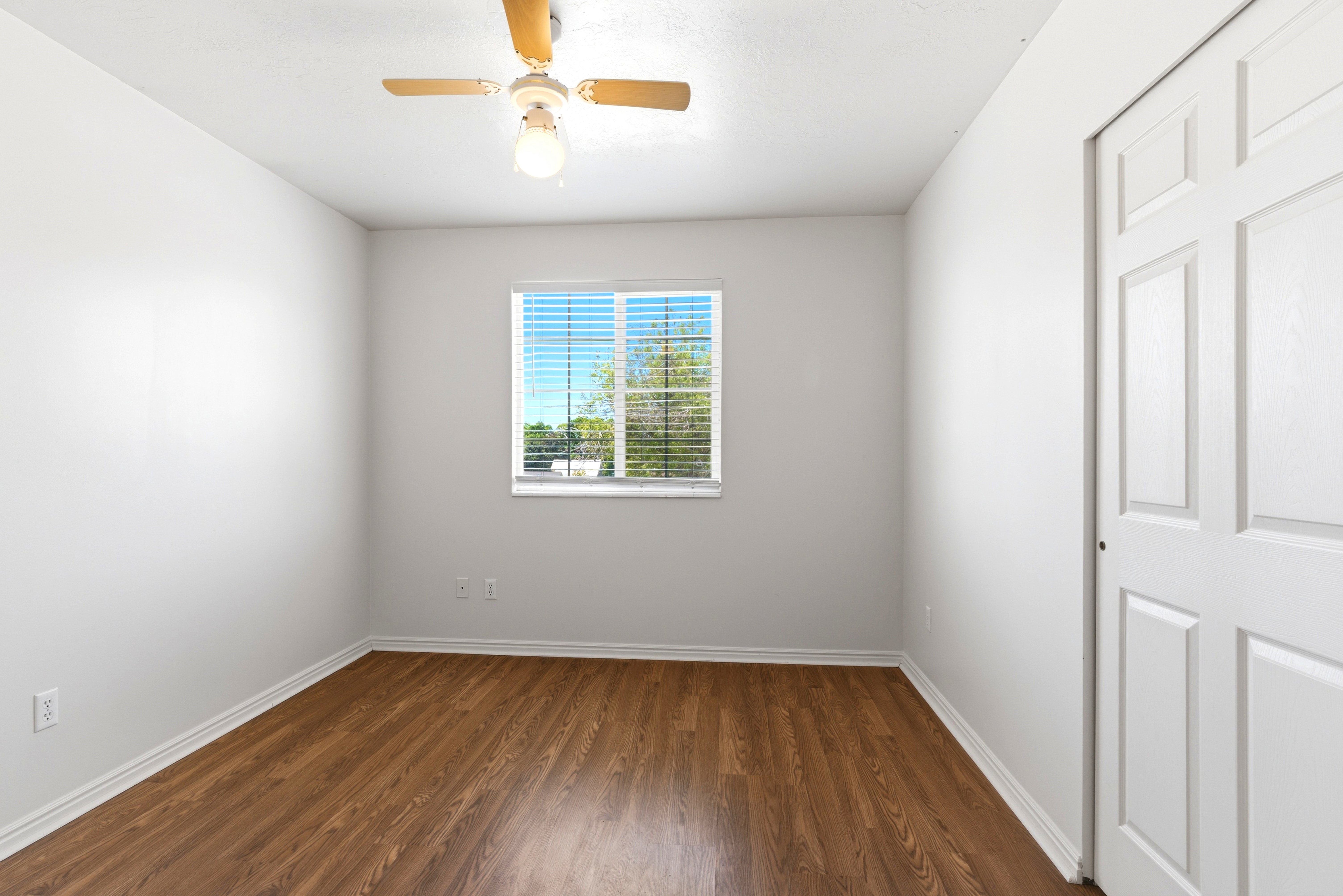 Bright interior space featuring wood-finish flooring, a window with blinds, a ceiling fan with integrated lighting, and a white bi-fold closet door