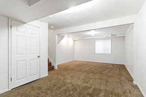 Expansive carpeted room featuring white walls, a white paneled door, wood-finish stairs, and a window with blinds