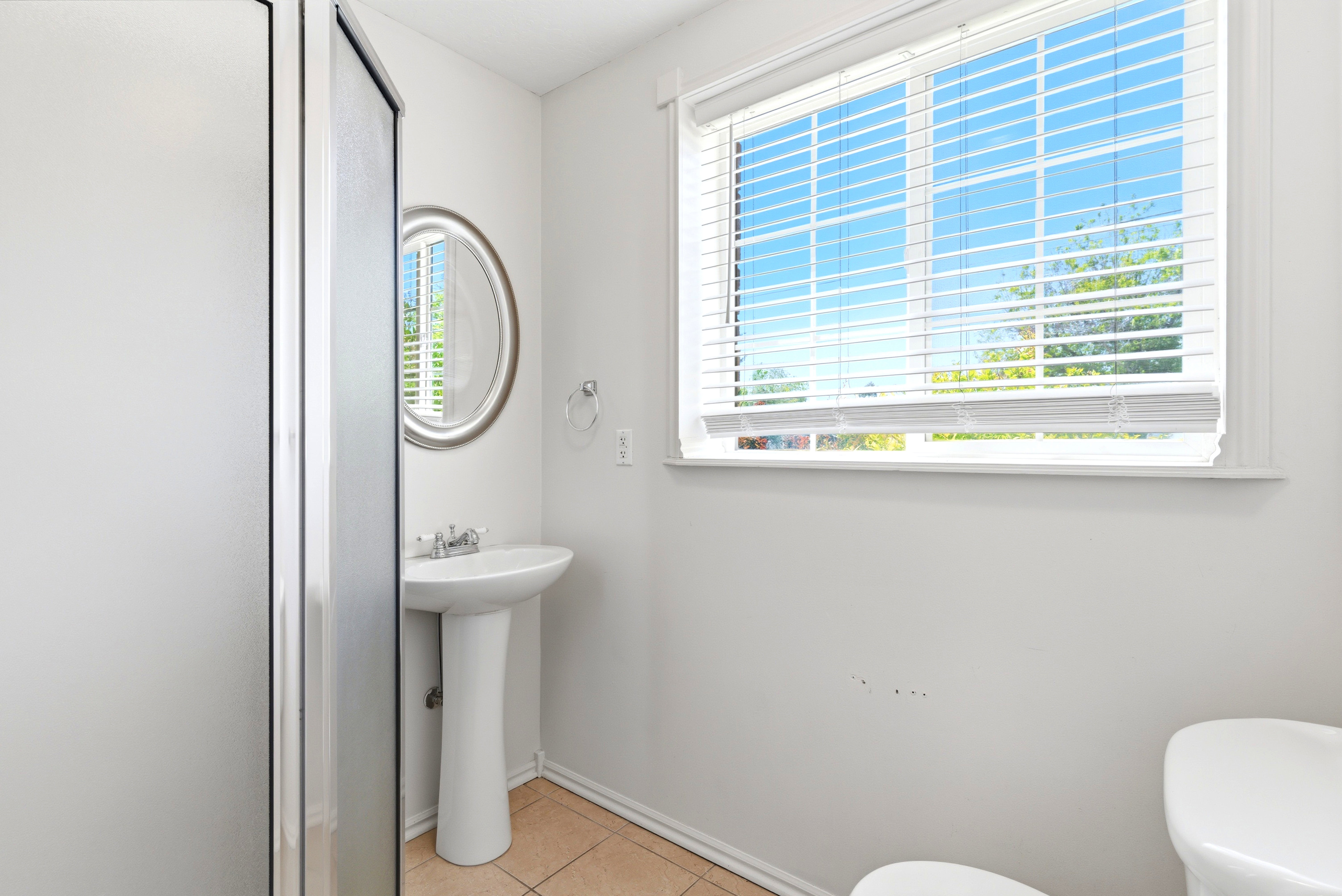 Bathroom featuring a pedestal sink, a framed oval mirror, a window with blinds, and tile flooring