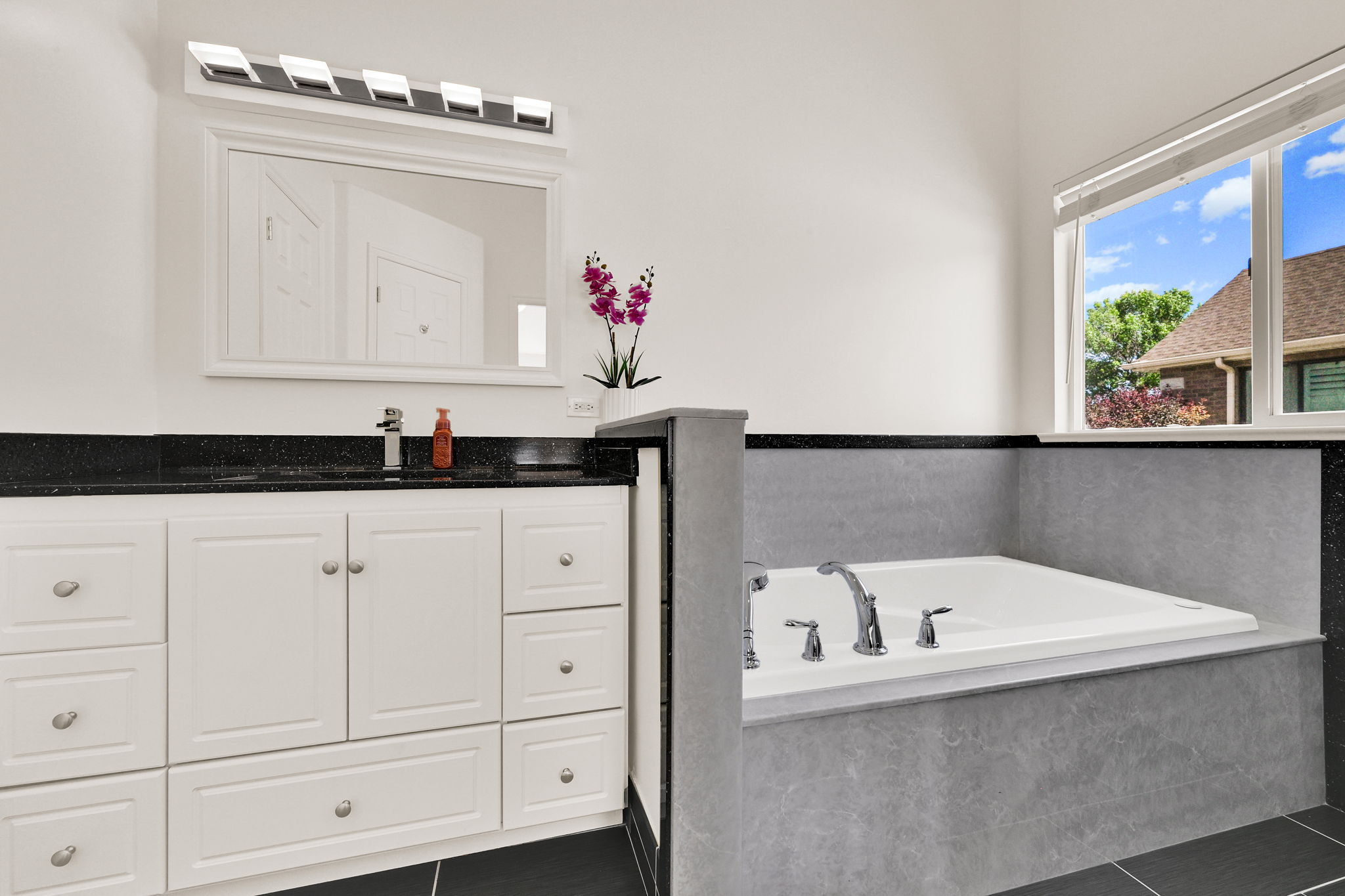 Built-in soaking tub with chrome fixtures, white vanity with black countertop, and a large mirror with overhead lighting
