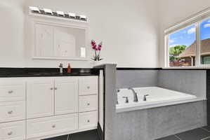 Built-in soaking tub with chrome fixtures, white vanity with black countertop, and a large mirror with overhead lighting