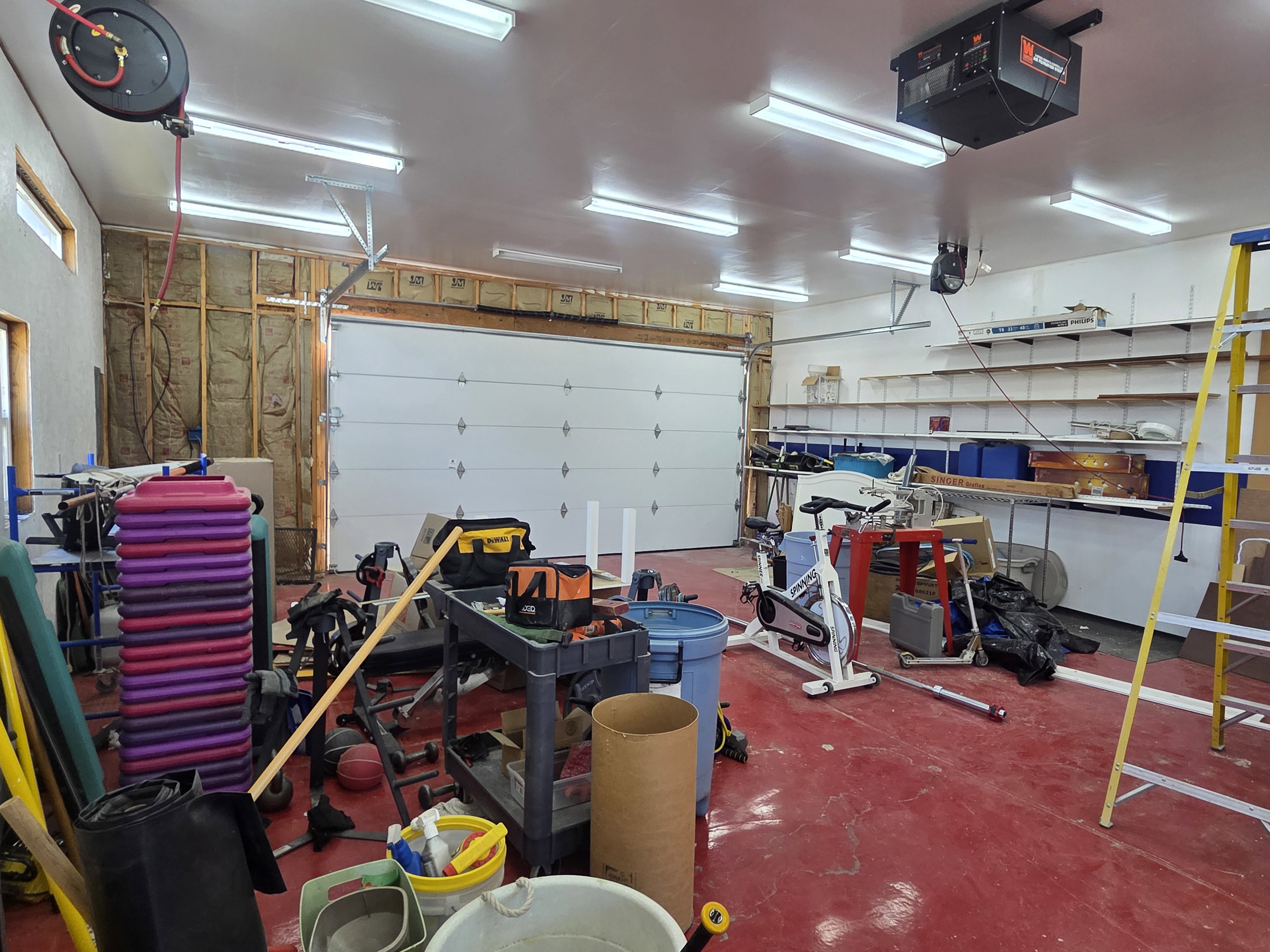 Spacious garage featuring a sectional garage door, multiple fluorescent ceiling lights, built-in wall shelving, and a red-painted concrete floor