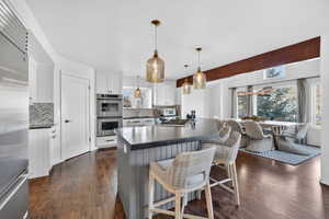 Modern kitchen featuring a large center island with seating, stainless steel double ovens, wood-finish flooring, white cabinetry, and a patterned tile backsplash
