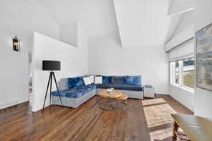 Spacious living area featuring high vaulted ceilings, rich wood-finish flooring, a large window with roller shades, a partial wall partition, and a wall-mounted sconce