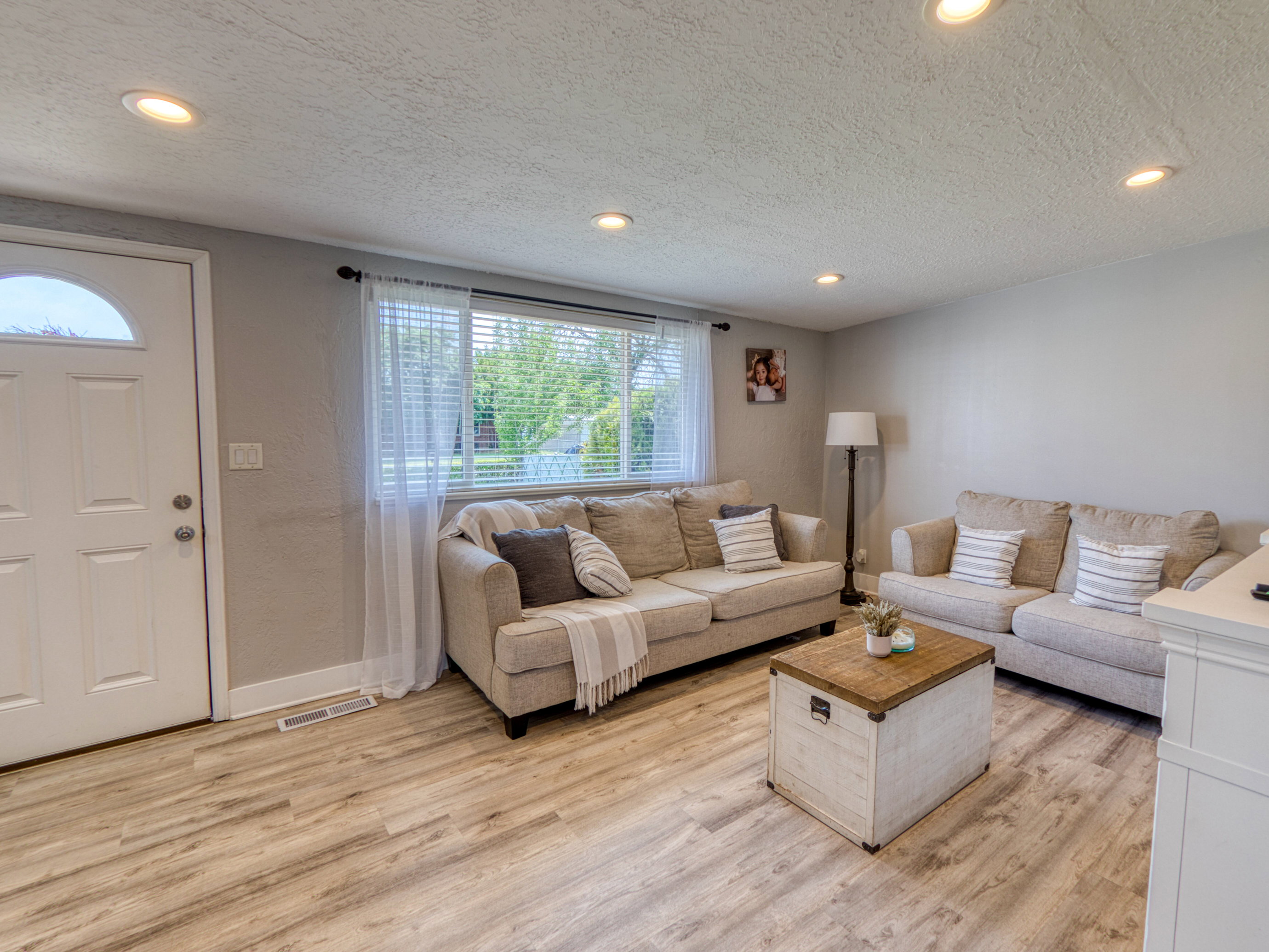 Living area with light wood-finish flooring, recessed lighting, a large window, and a white paneled entry door with an arched glass insert