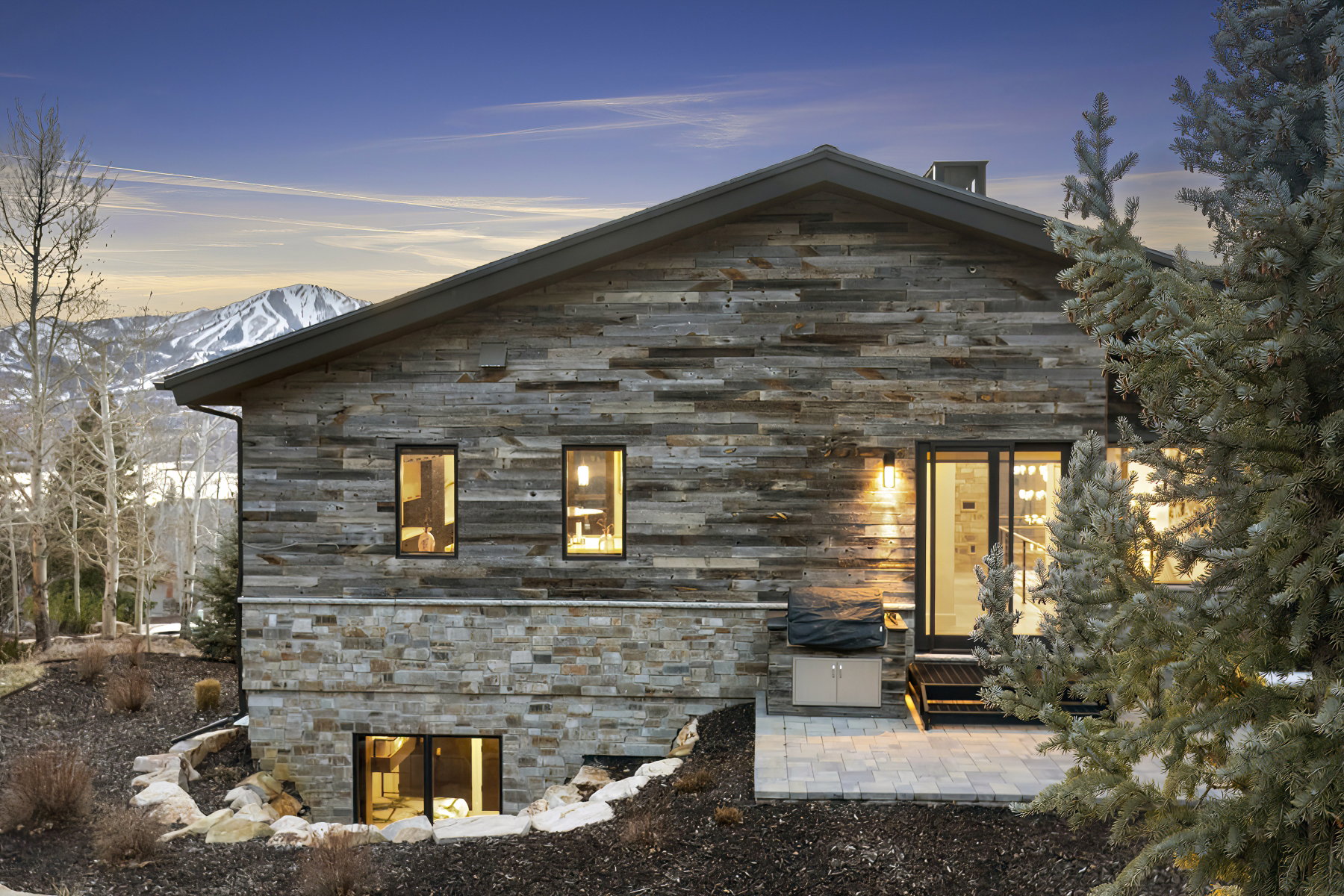 Exterior featuring horizontal wood siding, natural stone veneer, a sloped roofline, a ground-level patio with pavers, and a prominent mountain backdrop