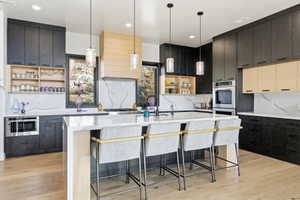 Modern kitchen featuring dark wood-finish cabinetry, light wood-finish accents, a waterfall island with seating, integrated stainless steel appliances, and white stone countertops with a matching backsplash