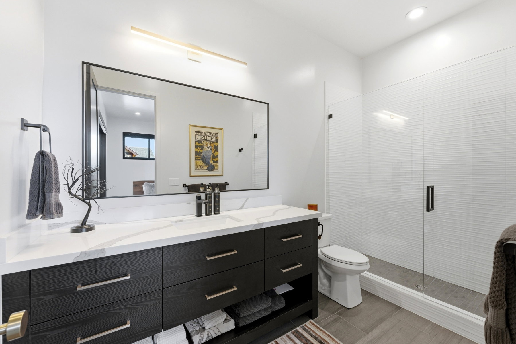 Spacious bathroom featuring a dark wood-finish vanity with a light-toned countertop, a large framed mirror with integrated lighting, a walk-in shower with clear glass enclosure, and textured white shower wall tiling