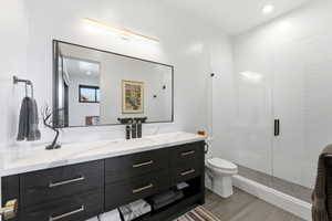 Spacious bathroom featuring a dark wood-finish vanity with a light-toned countertop, a large framed mirror with integrated lighting, a walk-in shower with clear glass enclosure, and textured white shower wall tiling