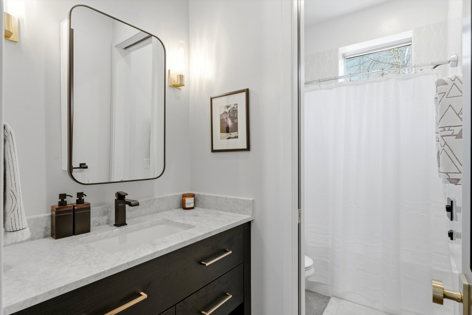 Modern bathroom featuring a dark wood-finish vanity with brass hardware, a white countertop with an integrated sink, a rectangular framed mirror, and wall-mounted sconces