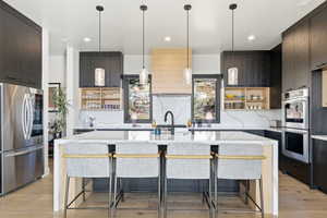 Gourmet kitchen featuring a waterfall island with integrated seating, stone countertops with a dramatic veining pattern, dark wood-finish cabinetry, stainless steel appliances, and a light wood-finish range hood
