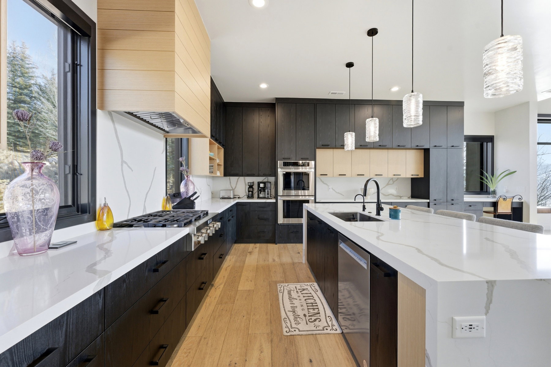 Modern kitchen featuring wood-finish flooring, two-tone cabinetry, a gas range with a wood-clad hood, large center island with integrated sink, and white countertops with veining