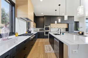 Modern kitchen featuring wood-finish flooring, two-tone cabinetry, a gas range with a wood-clad hood, large center island with integrated sink, and white countertops with veining
