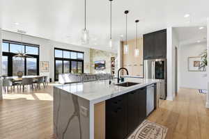 Open-concept kitchen featuring an oversized island with a waterfall countertop and integrated sink, stainless steel appliances, pendant lighting, and light wood-finish flooring