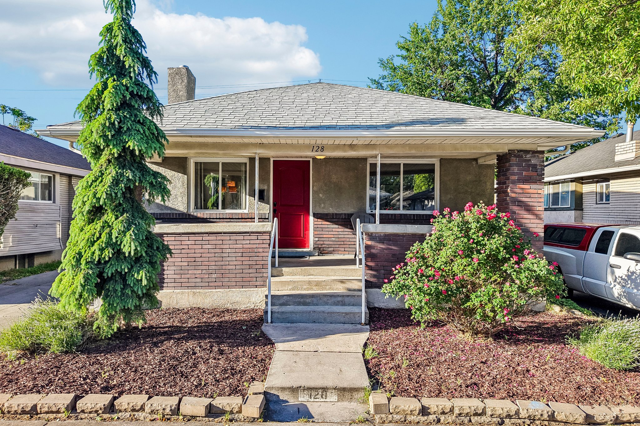 Front exterior featuring a red entry door, brick accents, concrete porch, and a manicured landscape with mature trees and shrubs