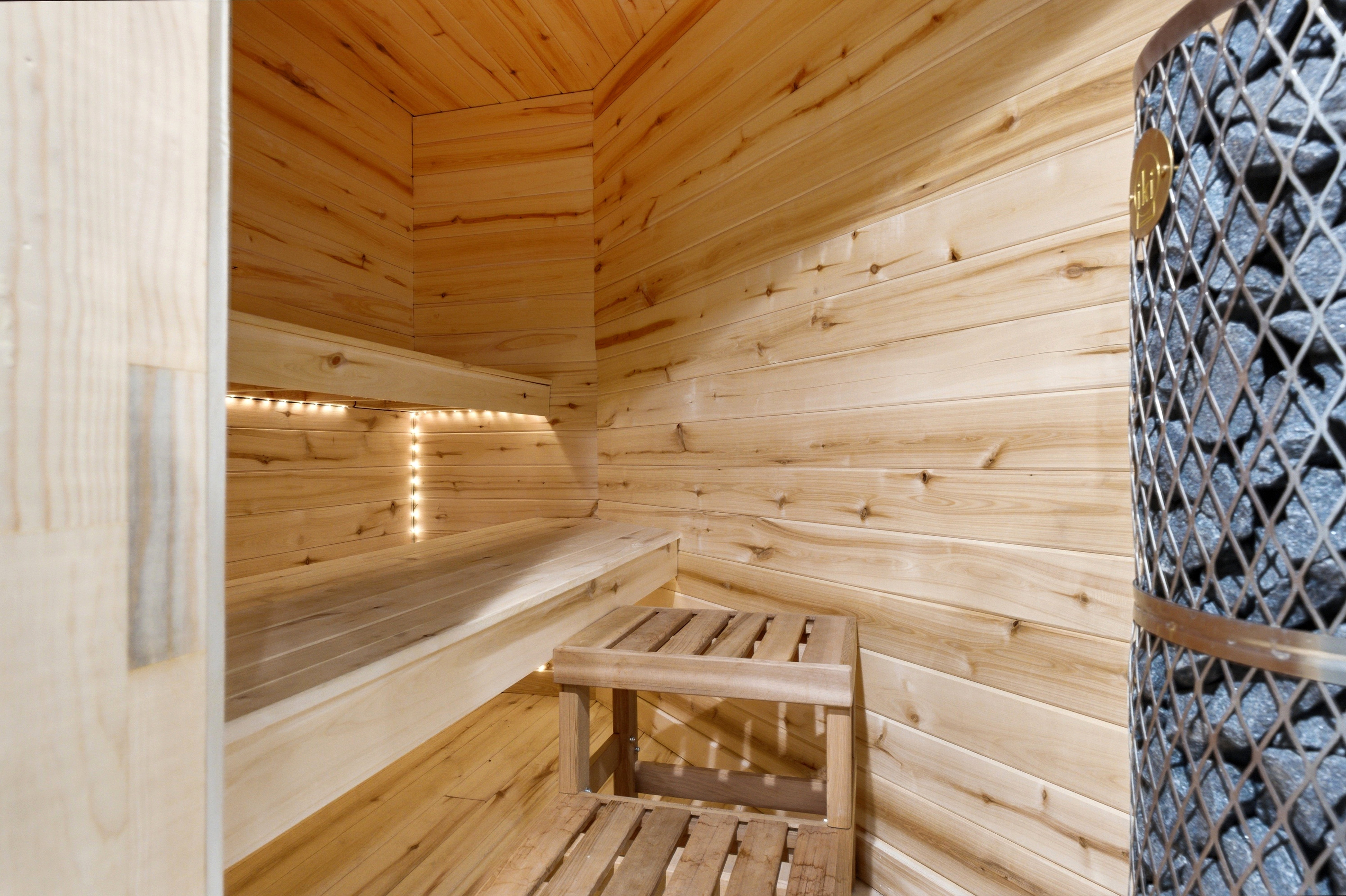 Wood-paneled sauna featuring a multi-tiered bench, integrated strip lighting, slatted wood flooring, and a stone heater with a metal cage