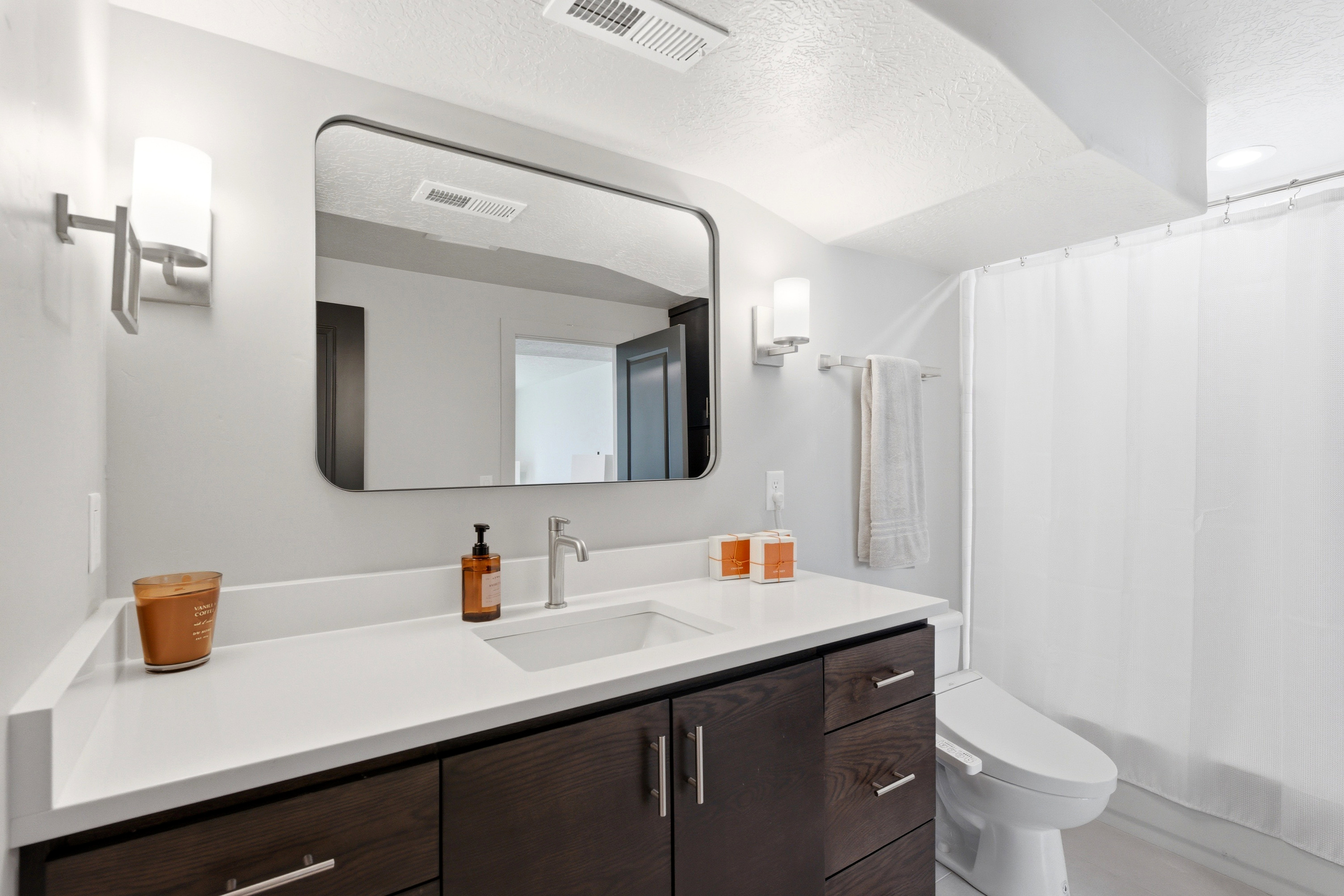 Bathroom featuring a dark wood-finish vanity with an integrated sink and white countertop, complemented by a rectangular frameless mirror and modern wall sconces