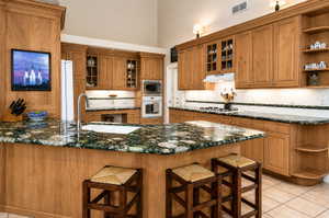 Kitchen featuring a central island with a built-in sink and granite-finish countertop, light wood cabinetry, white tile backsplash, and stainless steel appliances including a wall oven and microwave