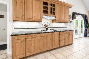 Kitchen featuring wood cabinetry, dark countertops, a white tile backsplash with decorative accents, and a built-in cooktop