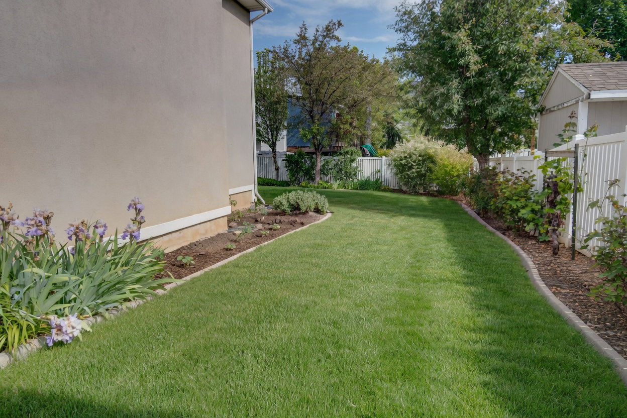 Expansive lawn area featuring a stucco exterior, a white vinyl fence, established perimeter landscaping, and mature trees