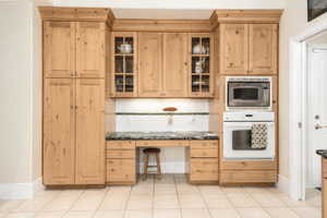 Built-in kitchen cabinetry featuring a natural wood finish, glass-front upper cabinets, integrated desk space, wall oven, and microwave